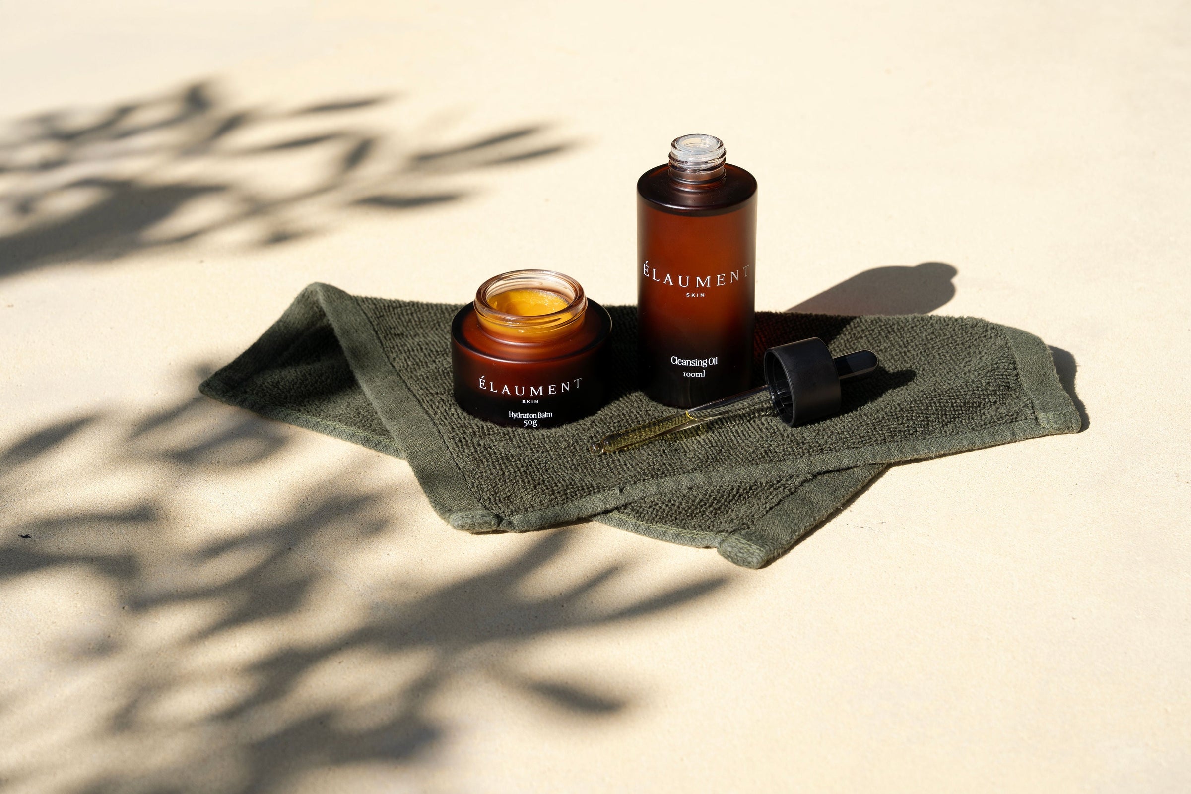 Élaument Skin Cleansing Oil and Hydration Balm on an olive towel in sunlight