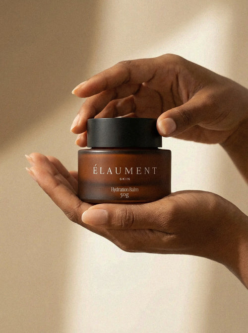 Hand holding a brown jar of Élaument skincare product against a beige background