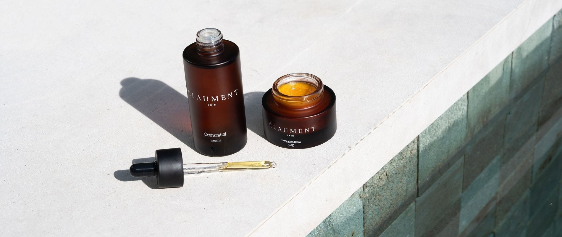 The Élaument Cleansing Oil and Hydration Balm on a stone ledge beside water